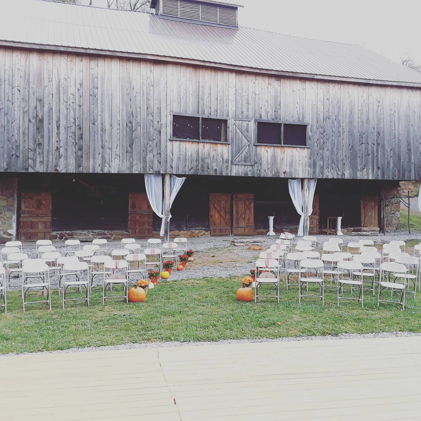 Weathered gray historic barn set up for a fall wedding ceremony with rows of white folding chairs and small pumpkins.