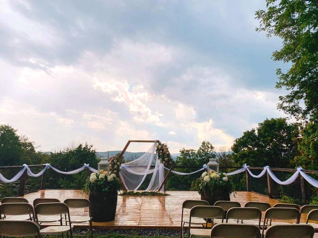 Outdoor wedding deck decorated with a floral hexagonal arch and white drapery, rows of empty chairs, set against a dramatic cloudy sky and mountain view.